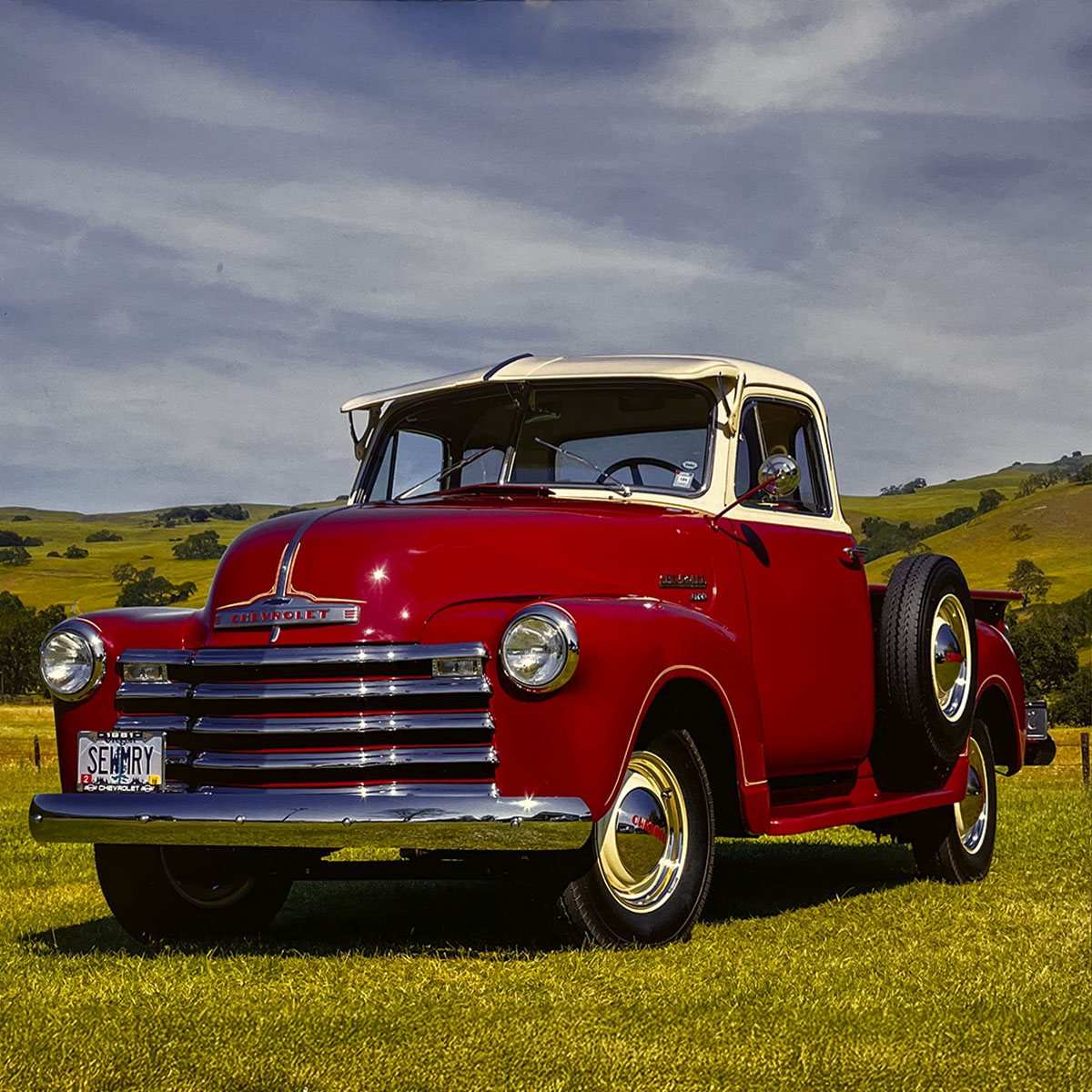 A striking red and cream two-tone 1951 Chevrolet 3100 pickup truck, parked in a lush green field under a partly cloudy sky, featuring a side-mounted spare tire and classic styling.