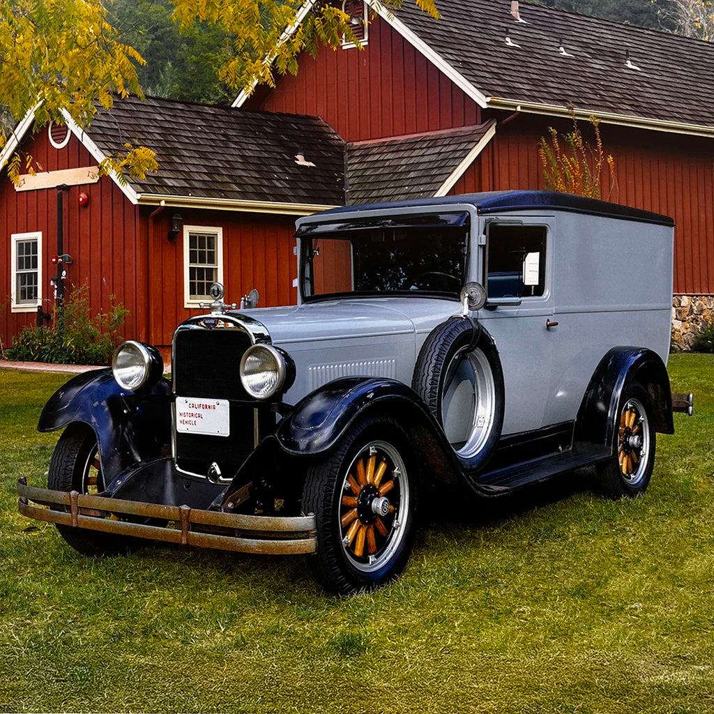 A distinguished grey and black 1929 Ford Model A Sedan Delivery van, parked on a green lawn in front of a rustic red barn, showcasing its vintage commercial vehicle design.