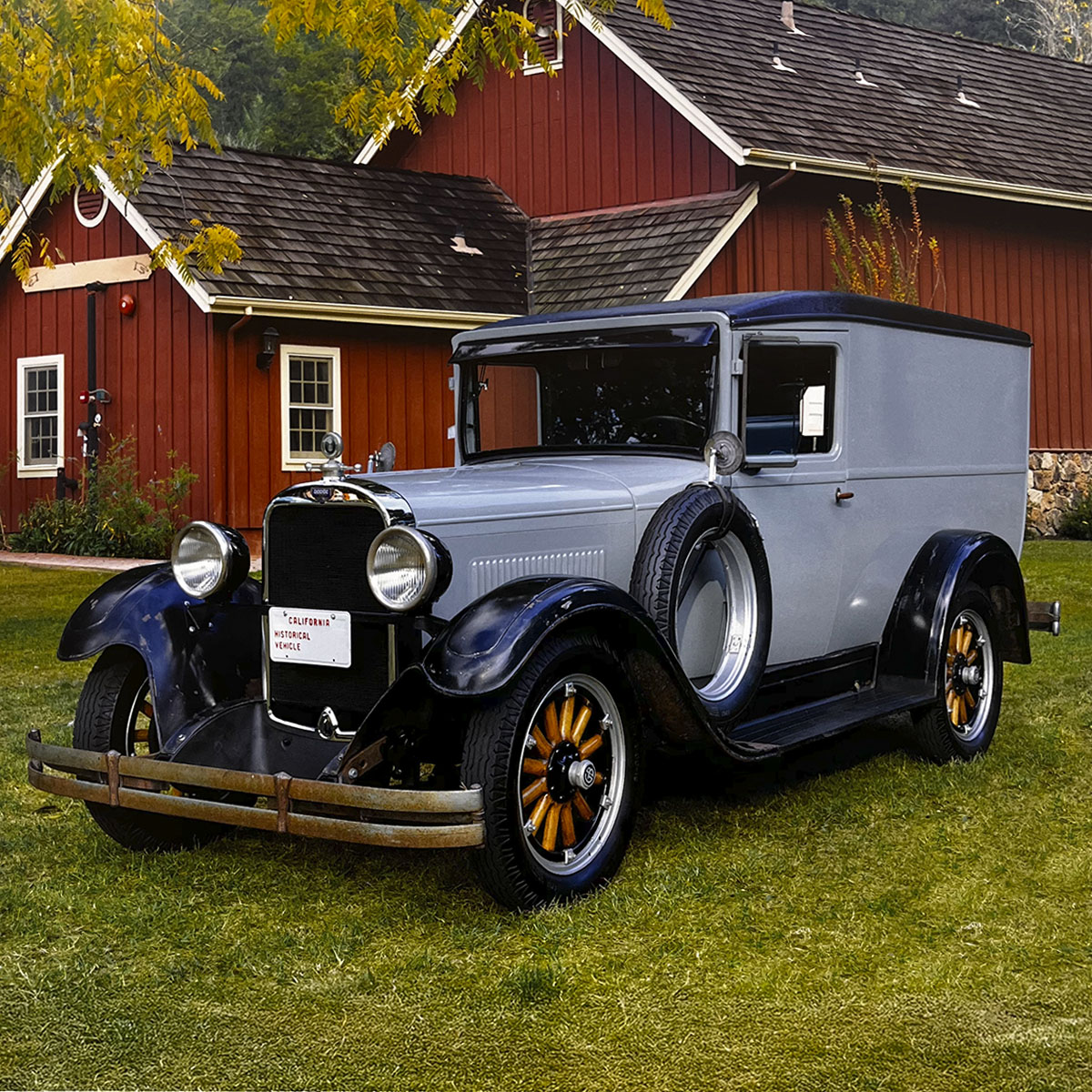 A distinguished grey and black 1929 Ford Model A Sedan Delivery van, parked on a green lawn in front of a rustic red barn, showcasing its vintage commercial vehicle design.