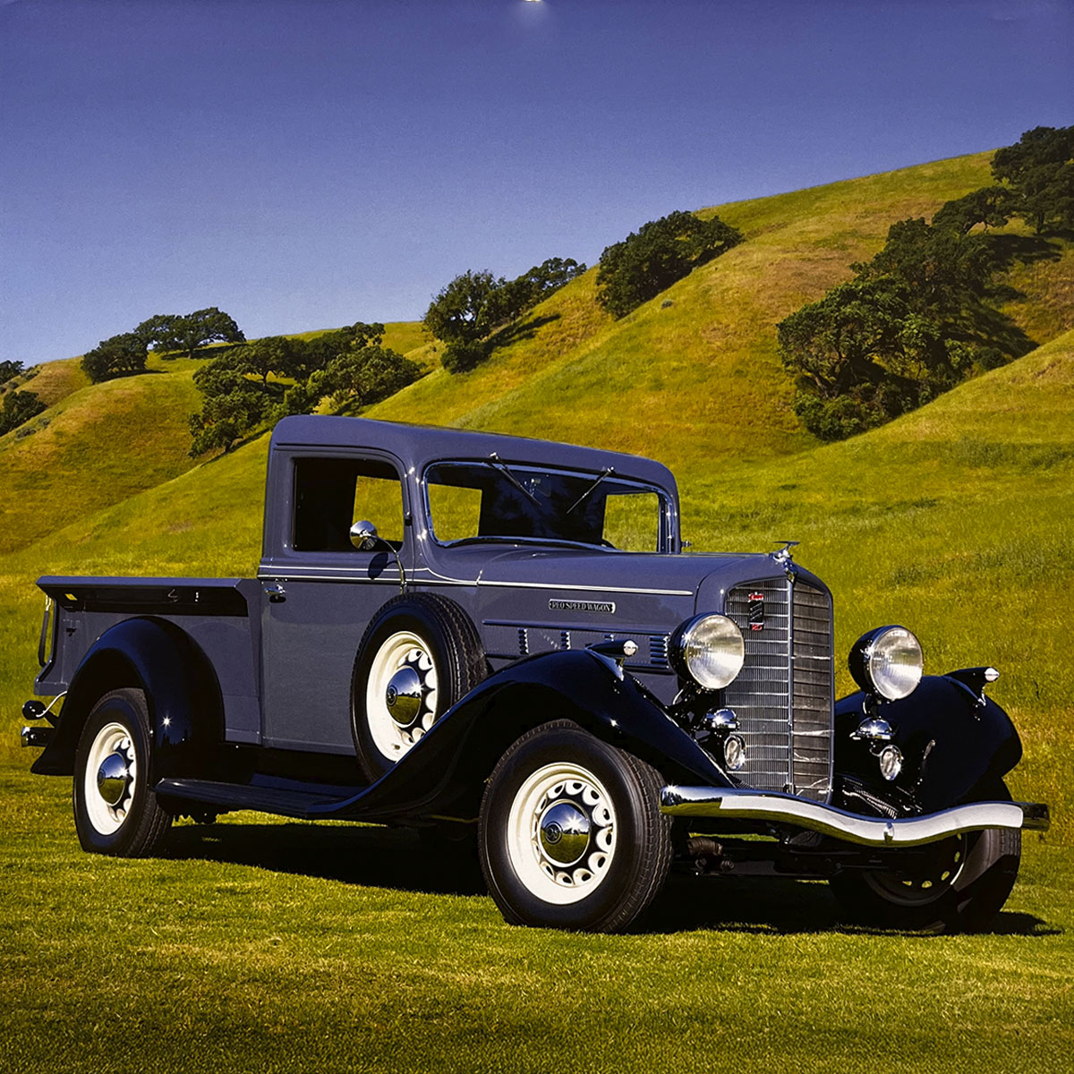 A stately dark grey 1937 International C-1 pickup truck with black fenders and white wall tires, resting on a verdant grassy hill against a bright blue sky, a testament to classic truck engineering.