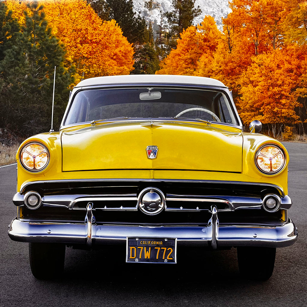 The iconic front view of a bright yellow 1953 Ford Crestline Victoria, featuring prominent chrome and round headlights, with a background of vibrant autumn trees, a classic American sedan.