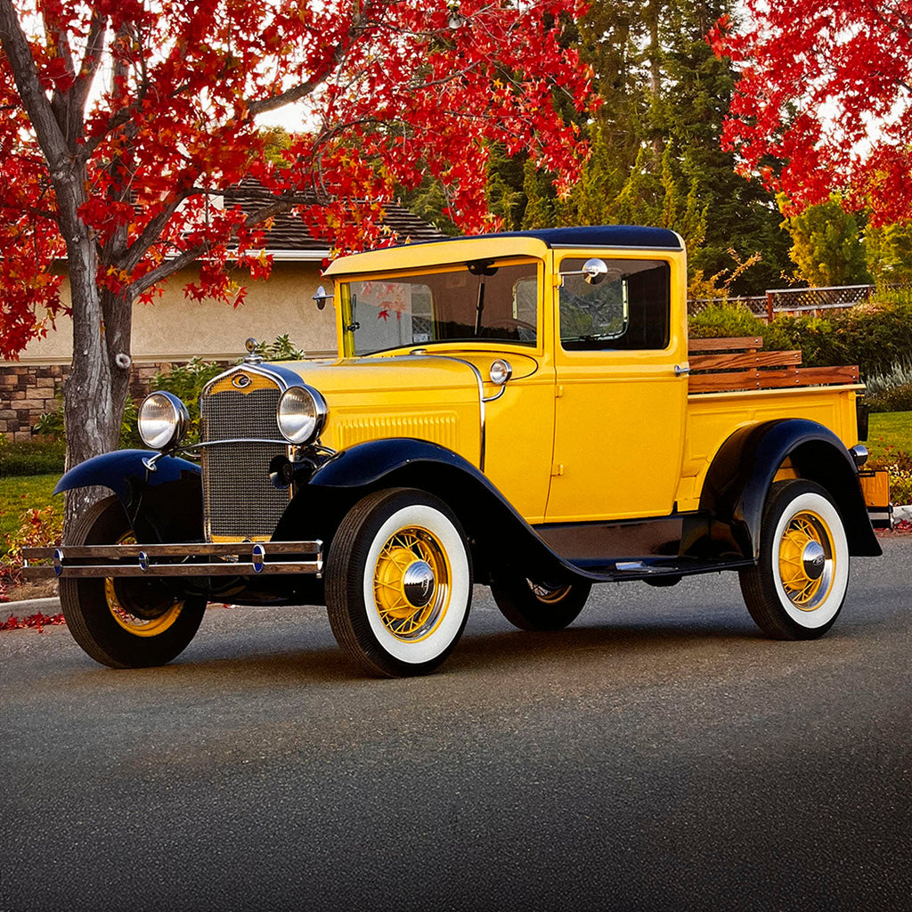 A brilliant yellow and black 1931 Ford Model A pickup truck with a wooden stake bed, parked on an asphalt road surrounded by colorful autumn foliage, celebrating vintage farm trucks.