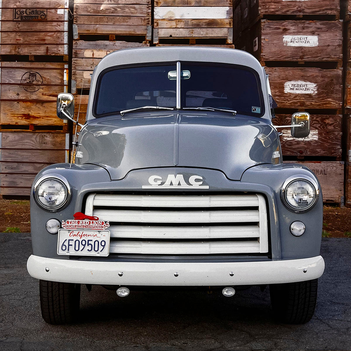 A sturdy grey 1949 GMC FC102 Series 100 pickup truck, presented from the front with a distinctive white grille and bumper, against a backdrop of stacked wooden crates, a symbol of post-war utility.