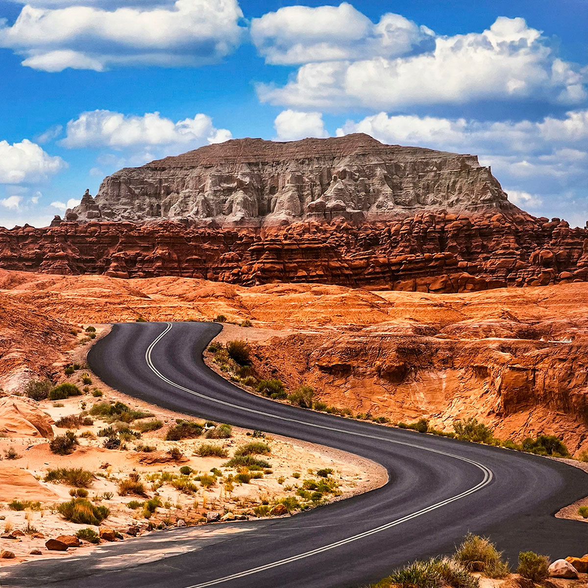 Winding paved road through a vast red rock desert landscape under a dramatic blue sky with white clouds, highlighting the beauty of arid natural terrain.