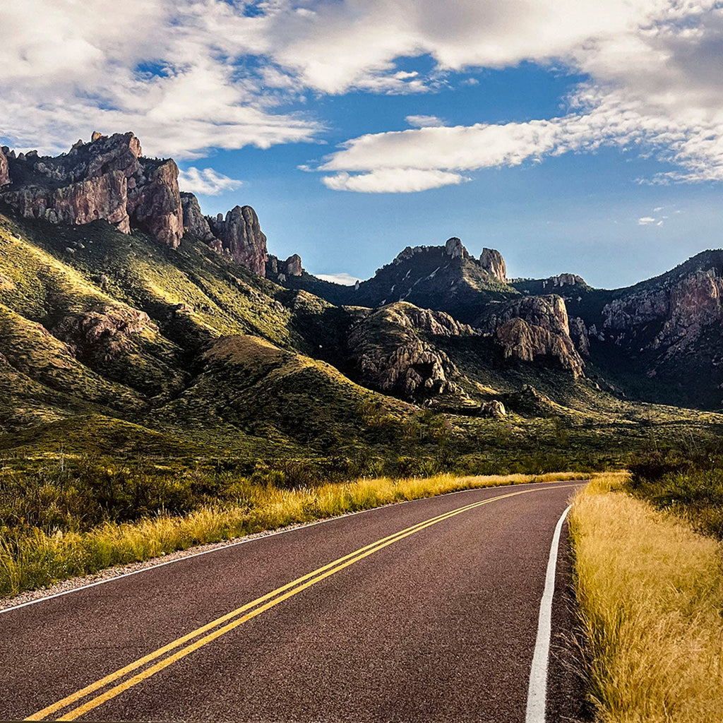 Scenic two-lane road winding through rolling green hills and rugged, sun-drenched mountains under a bright blue sky, perfect for a picturesque drive.