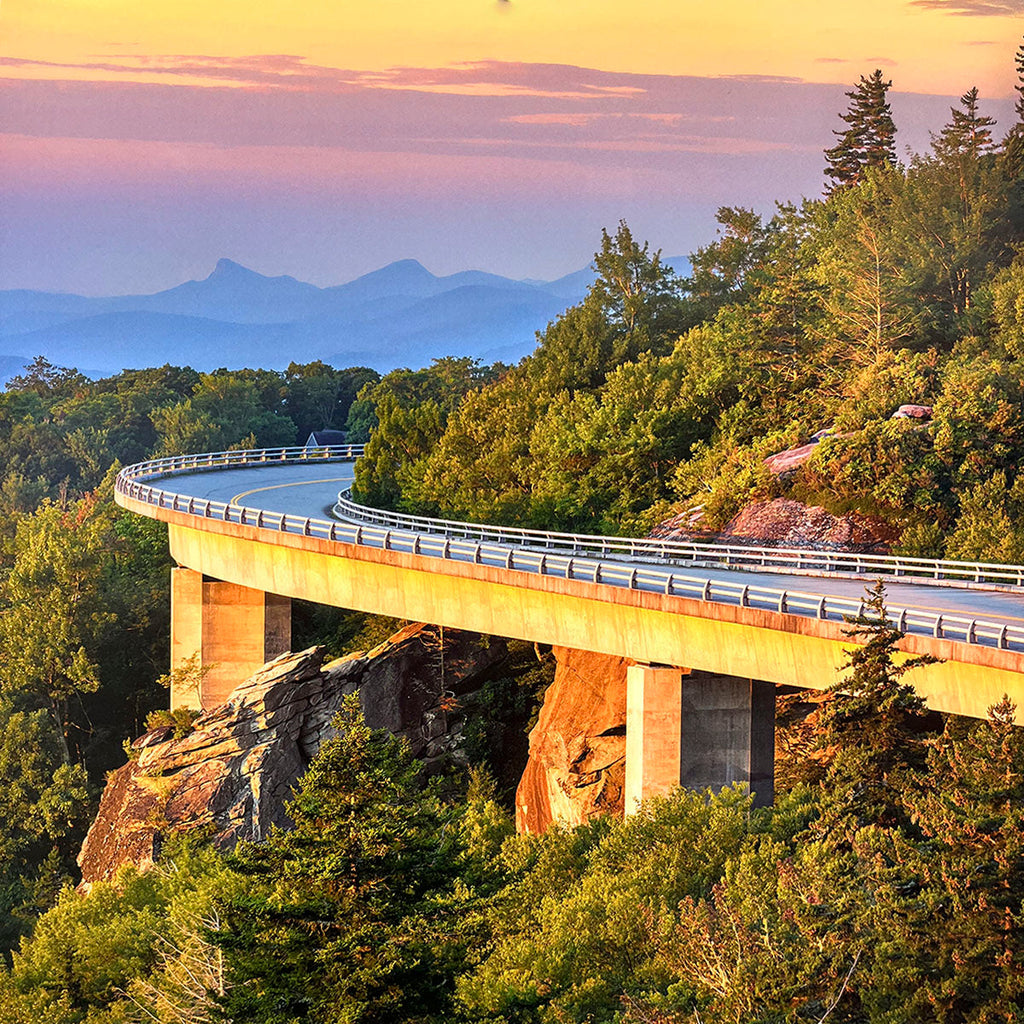 Elevated highway bridge curving gracefully around a lush, tree-covered mountain, illuminated by the warm glow of a sunset sky, offering a panoramic view.