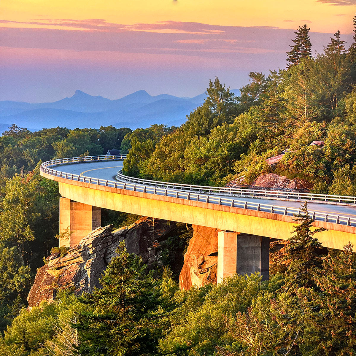 Elevated highway bridge curving gracefully around a lush, tree-covered mountain, illuminated by the warm glow of a sunset sky, offering a panoramic view.