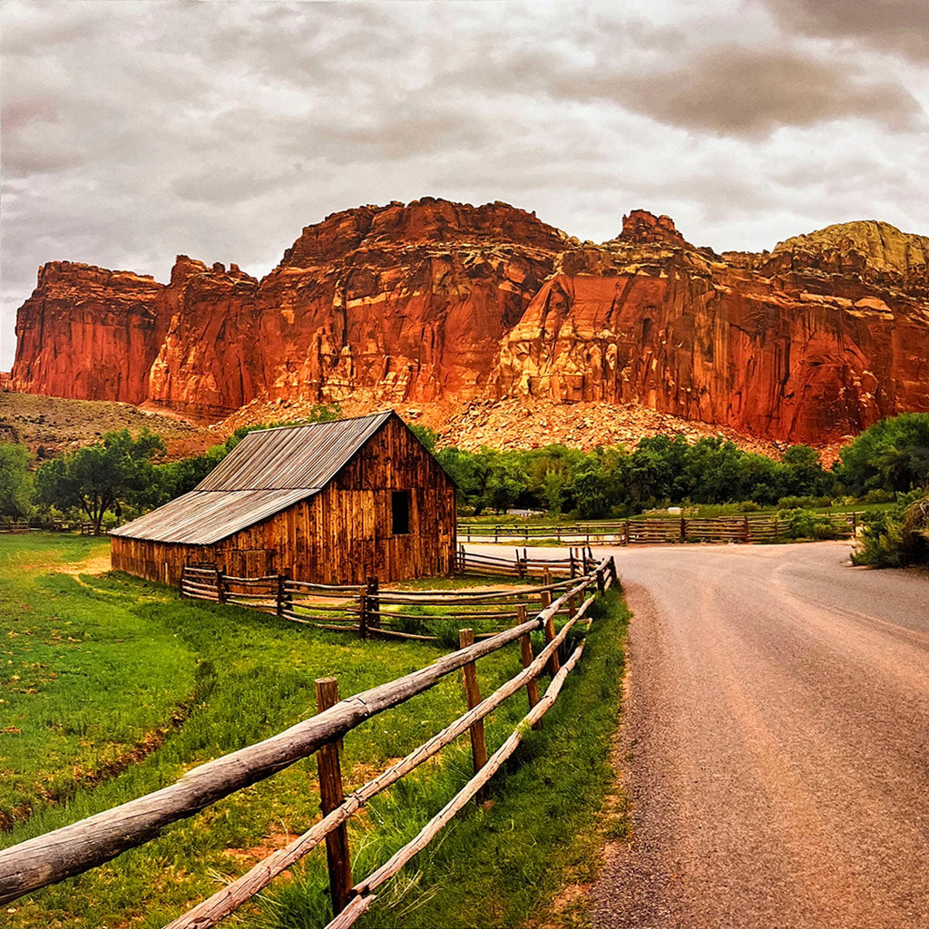 Rustic wooden barn with a split-rail fence next to a dirt road, set against a backdrop of imposing red rock cliffs under a dramatic cloudy sky, depicting a pastoral scene.