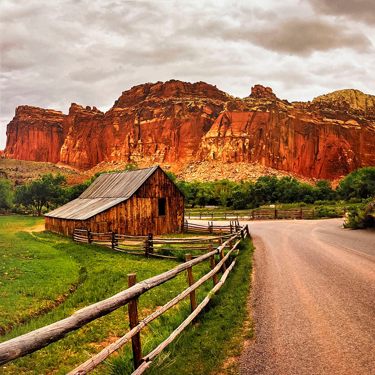 Rustic wooden barn with a split-rail fence next to a dirt road, set against a backdrop of imposing red rock cliffs under a dramatic cloudy sky, depicting a pastoral scene.