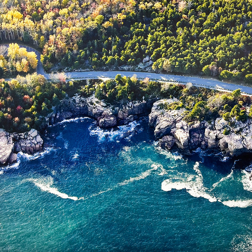 Aerial view of a coastal road hugging a rugged, rocky shoreline with deep blue ocean waves crashing against the cliffs, highlighting a dramatic seascape.