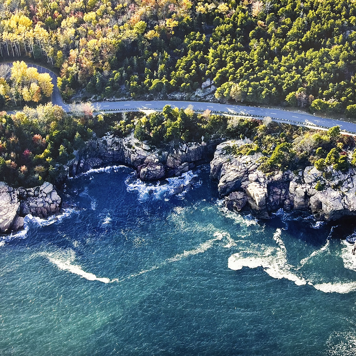Aerial view of a coastal road hugging a rugged, rocky shoreline with deep blue ocean waves crashing against the cliffs, highlighting a dramatic seascape.
