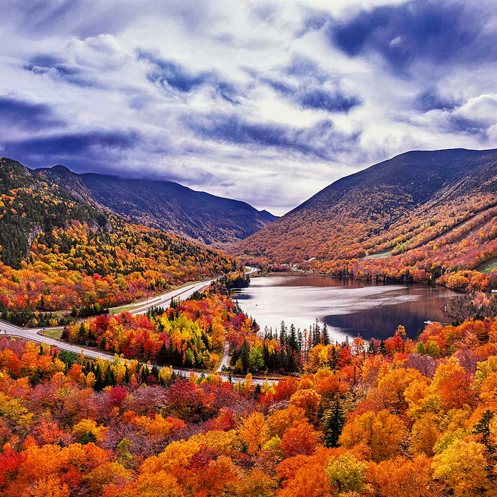 Breathtaking aerial view of a valley in full autumn splendor, with a serene lake reflecting vibrant orange, red, and yellow trees under a cloudy sky, showcasing peak fall foliage.