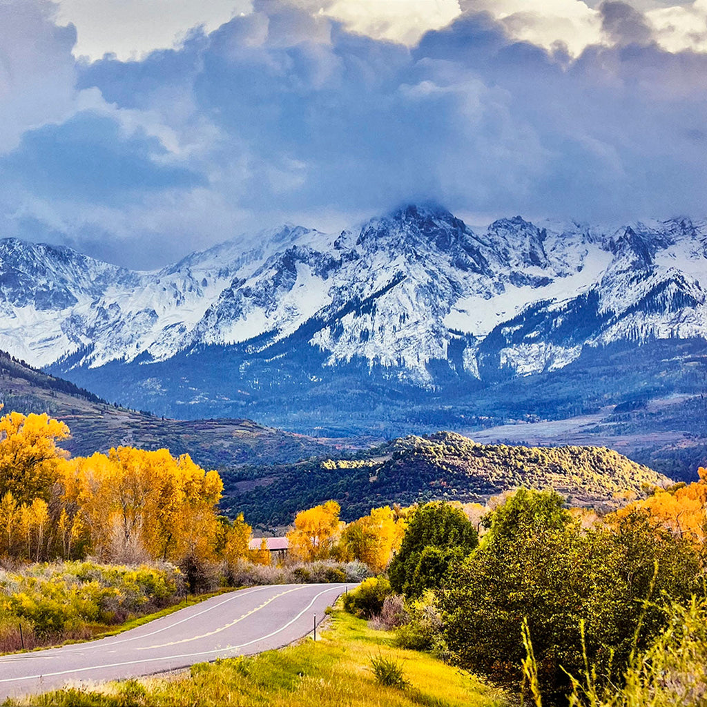 Scenic road winding through golden autumn trees with majestic snow-capped mountains towering in the background under a dramatic cloudy sky, illustrating seasonal contrasts.