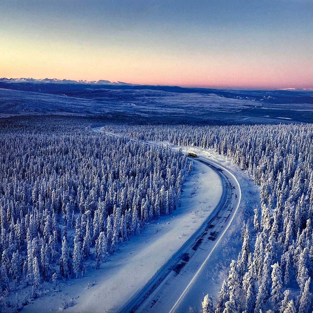Aerial view of a lone vehicle on a snowy road cutting through an expansive forest of snow-covered pine trees during a soft twilight or sunrise, capturing a serene winter scene.