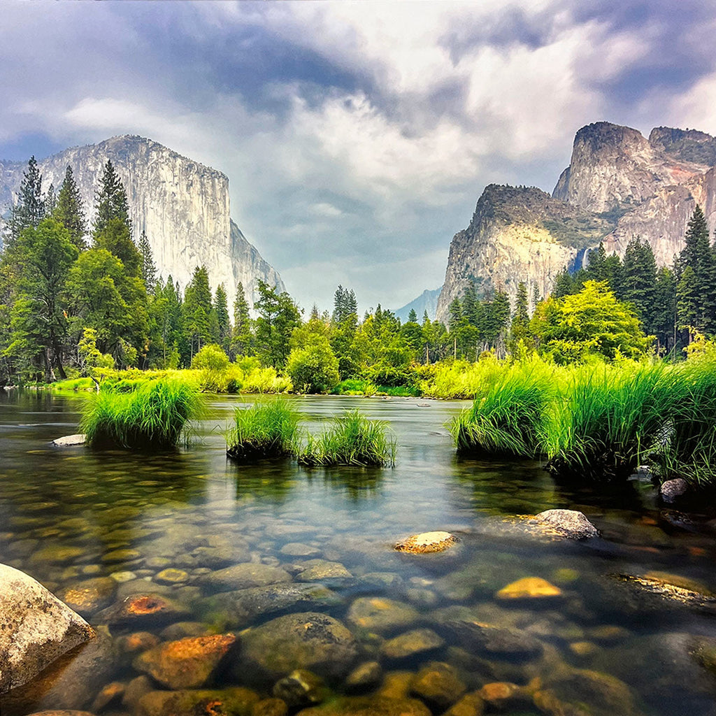 Serene Yosemite-style valley landscape for wall calendar, featuring a clear, rocky river flowing through lush green banks and towering granite cliffs under a cloudy sky. SEO: river valley, green landscape, tall mountains, clear water, scenic calendar.