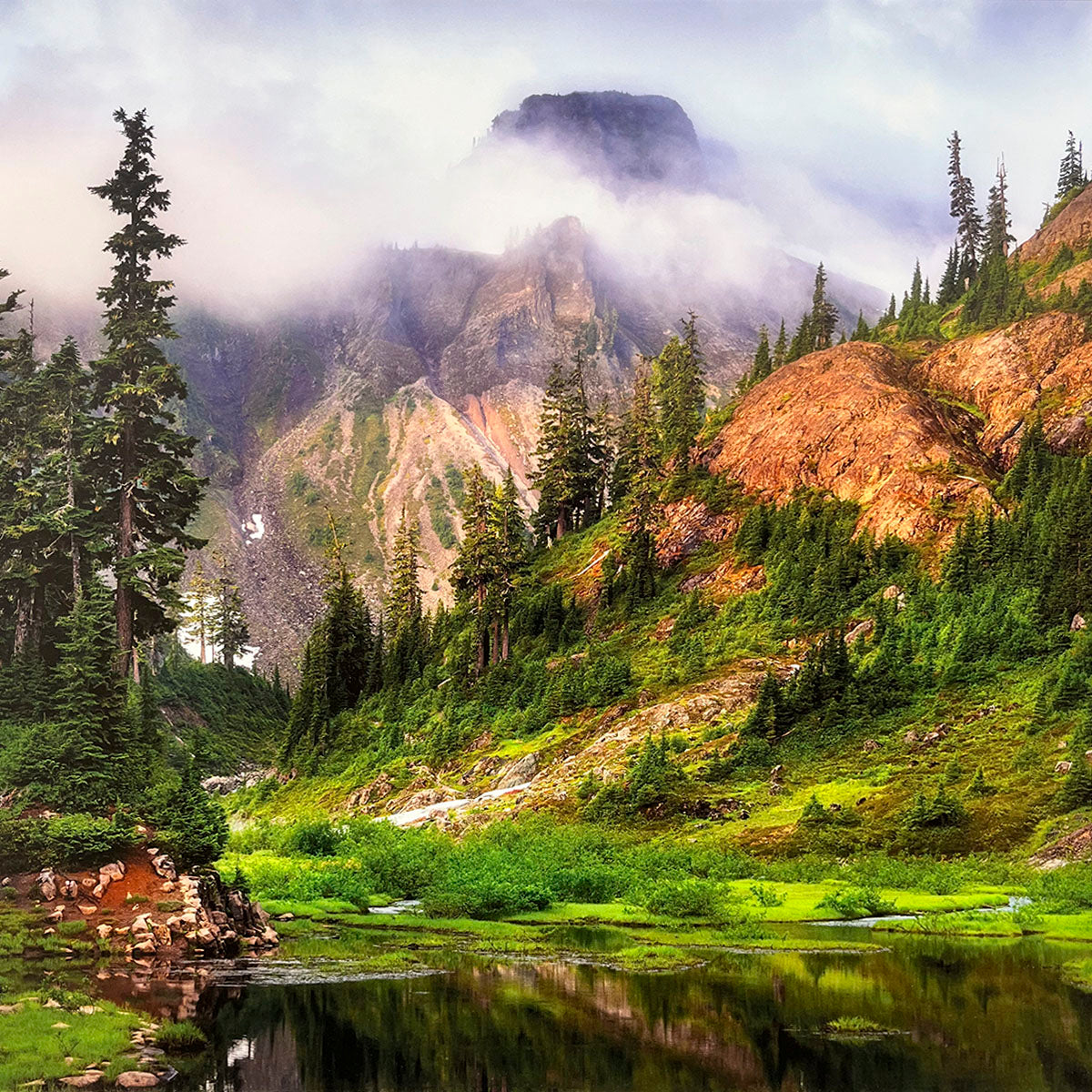 Misty mountain scene for wall calendar, depicting a tranquil dark lake reflecting towering peaks partially obscured by fog, surrounded by lush green forests and meadows. SEO: misty mountains, serene lake, foggy landscape, green forest, scenic calendar.