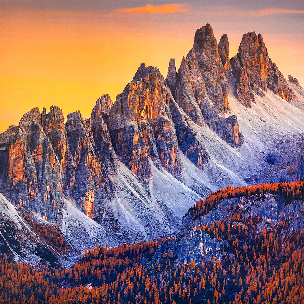 Jagged, snow-dusted mountain range for wall calendar, glowing with warm orange light from a sunset, with a dense forest of orange and red autumn trees below. SEO: jagged mountains, sunset glow, autumn forest, snow-dusted peaks, scenic wall calendar.