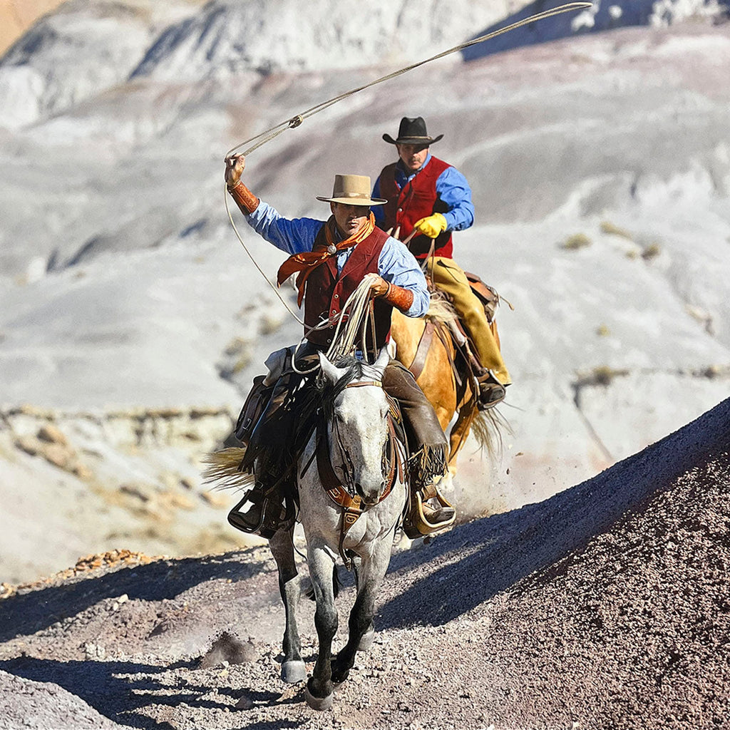 Two cowboys on horseback, one preparing to lasso, in a vast, arid mountainous landscape, highlighting Western ranching action for a calendar.