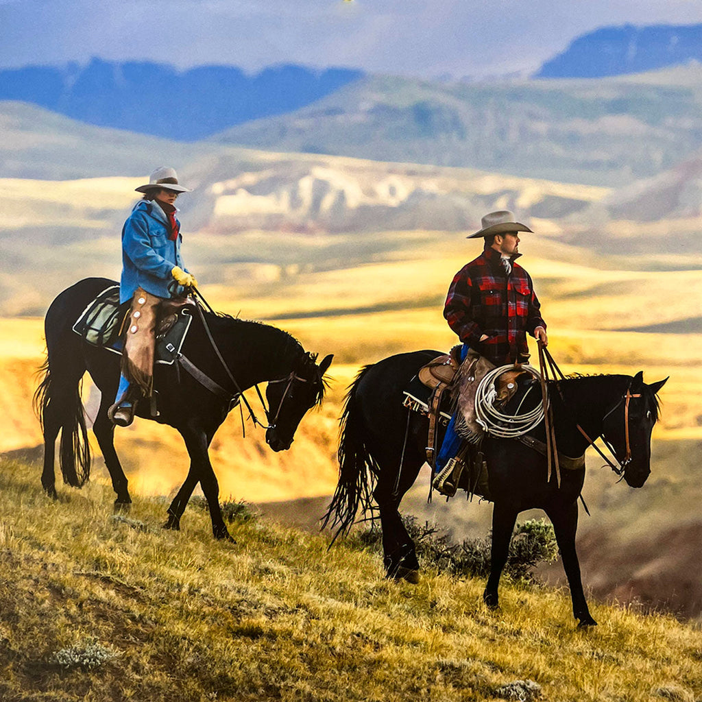 Two cowboys riding horses across a golden, hilly landscape at sunset, presenting a scenic and tranquil Western vista for a calendar.