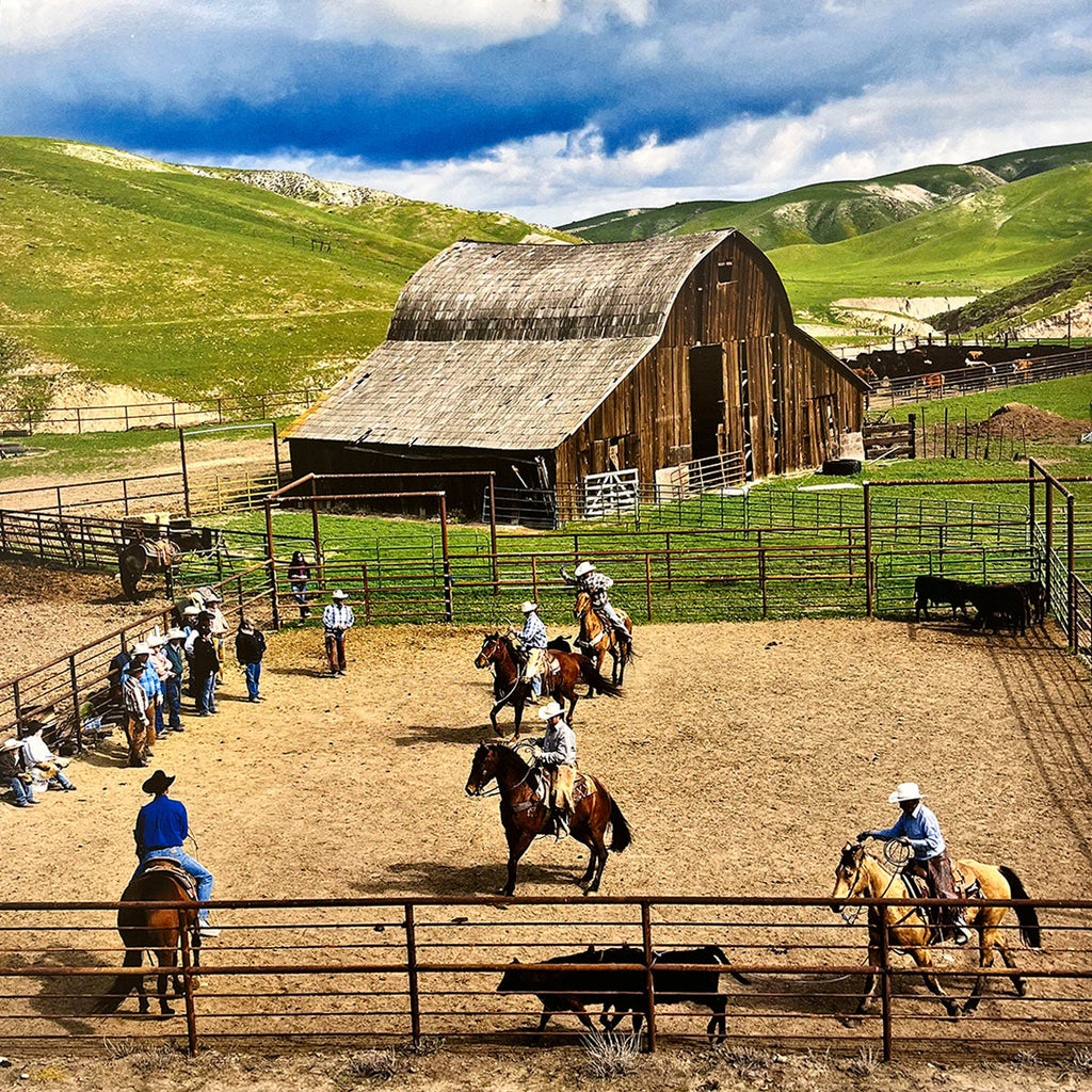High-angle view of a bustling rodeo or ranching arena with cowboys on horseback working cattle near a large wooden barn and green hills.