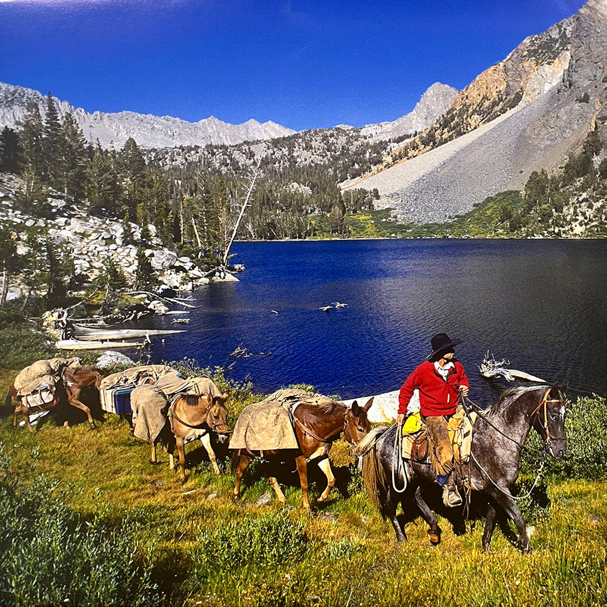 Cowboy on a horse leading a string of pack mules alongside a clear blue mountain lake with rugged peaks in the background, a Western adventure scene.