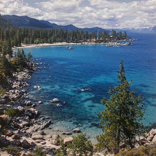 Stunning aerial view of Lake Tahoe's crystal-clear blue waters, rocky shorelines, lush green pine forests, and distant snow-capped mountains under a partly cloudy sky. Perfect for a landscape calendar, travel photography.