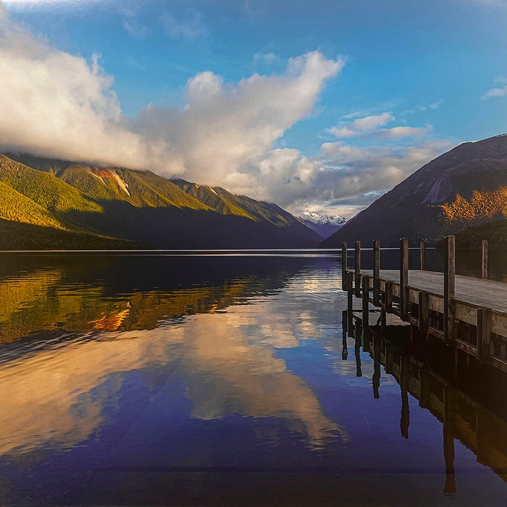 Picturesque lake scene with a rustic wooden dock extending into reflective blue waters, surrounded by majestic green mountains under a dramatic sky with white clouds. Keywords: mountain lake calendar, peaceful scenery, photography.