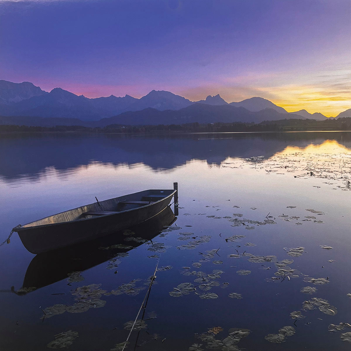 Serene sunset over a calm lake with a solitary wooden rowboat tied near lily pads, reflecting vibrant purple and orange sky colors, and framed by distant mountain silhouettes. Ideal for a tranquil nature calendar, lake art.