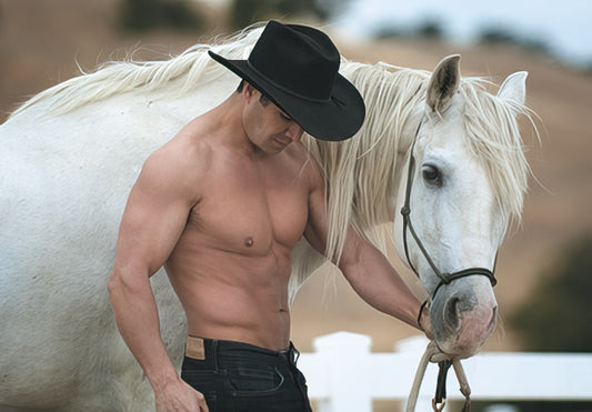 Handsome shirtless cowboy in a black hat with a muscular physique, gently holding the reins of a majestic white horse against a natural background.