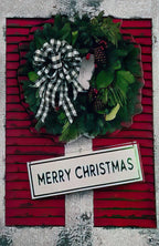 Front of a vibrant festive greeting card featuring a lush green wreath adorned with pinecones and a stylish black and white buffalo check bow, set against a rich red background with a 'MERRY CHRISTMAS' sign.