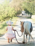 Young girl in a white cowboy hat and pink tutu, holding the reins of a gentle brown and white pony, walking down a sunlit dirt path on a rural farm. Perfect for cute child-friendly greeting cards.
