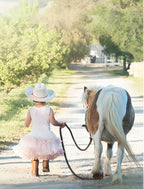 Young girl in a white cowboy hat and pink tutu, holding the reins of a gentle brown and white pony, walking down a sunlit dirt path on a rural farm. Perfect for cute child-friendly greeting cards.