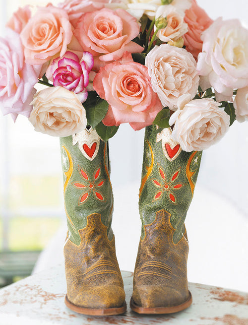 Vibrant bouquet of blush pink and white roses arranged artfully in rustic green and brown cowboy boots, perfect for a unique, lighthearted greeting card design.