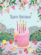 Front of a vibrant, whimsical greeting card featuring a multi-layered pink birthday cake with colorful candles, surrounded by strawberries, macarons, and decorative bunting. Small birds and butterflies flutter around, all against a pastel blue sky with green floral ground. The text 'Happy Birthday' is prominently displayed above the cake.