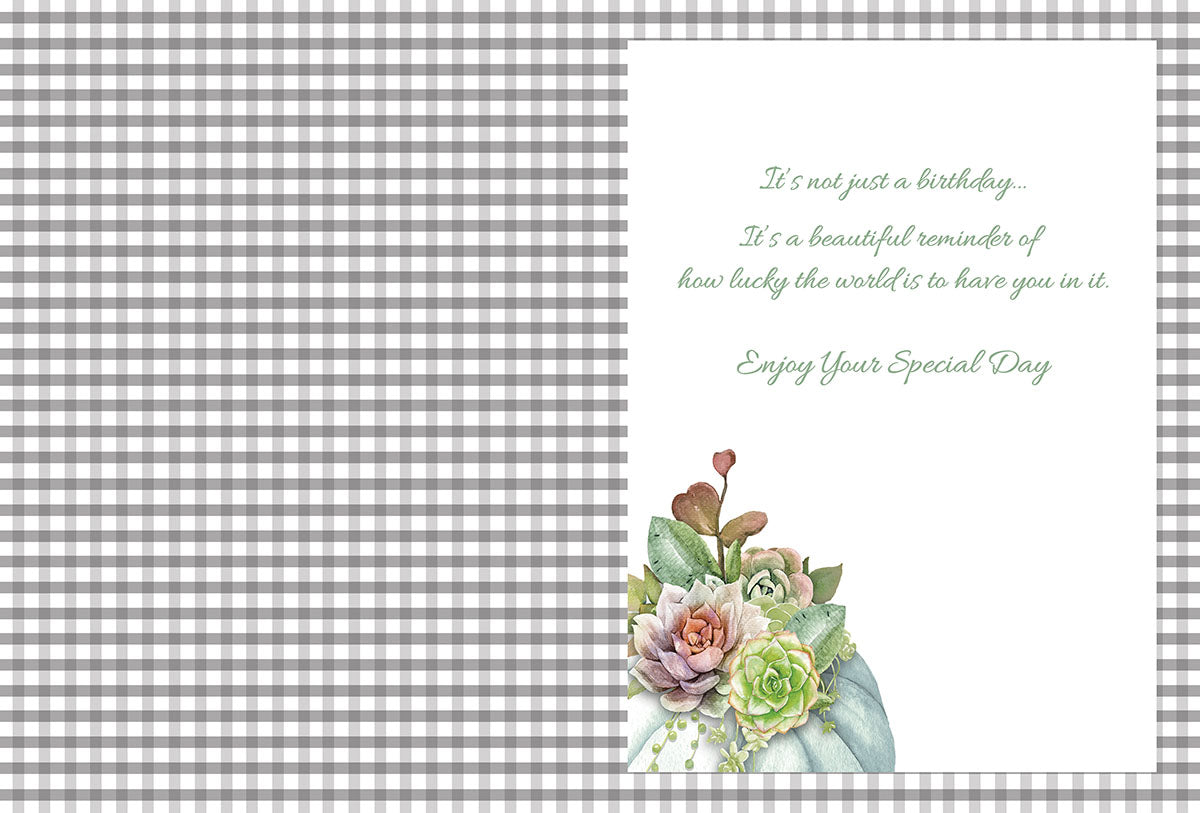 Open greeting card interior featuring a blank grey and white gingham pattern on the left. The right side displays a heartfelt script message: 'It's not just a birthday... It's a beautiful reminder of how lucky the world is to have you in it. Enjoy Your Special Day.' A small succulent and floral cluster adorns the bottom right corner.