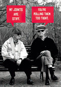 Humorous birthday card featuring two older men on a park bench, one smoking a cigarette, with speech bubbles reading 'MY JOINTS ARE STIFF.' and 'YOU'RE ROLLING THEM TOO TIGHT.' A funny adult card for him.