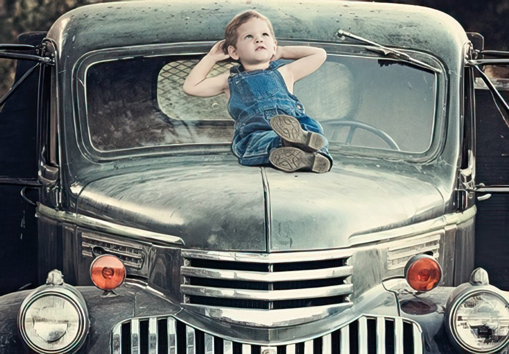 Cute young boy in denim overalls relaxing on the hood of a classic vintage green pickup truck, looking towards the sky, perfect for a child's birthday card.
