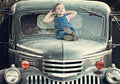Cute young boy in denim overalls relaxing on the hood of a classic vintage green pickup truck, looking towards the sky, perfect for a child's birthday card.