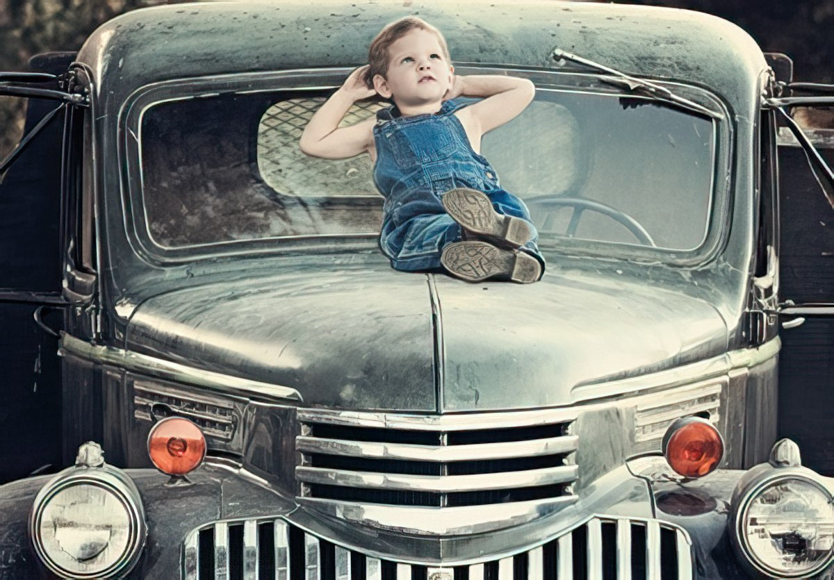 Cute young boy in denim overalls relaxing on the hood of a classic vintage green pickup truck, looking towards the sky, perfect for a child's birthday card.