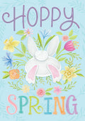 Cute Easter card front featuring a fluffy white bunny tail and pink feet emerging from vibrant spring flowers like daffodils, tulips, and cornflowers, with 'HOPPY SPRING' text on a light blue background.