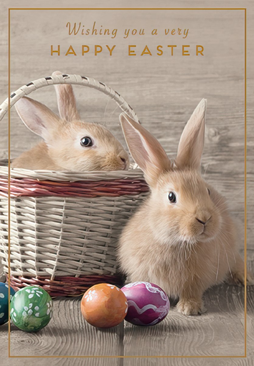 Front view of an adorable Easter greeting card featuring two fluffy brown bunnies, one peeking from a wicker basket and another sitting beside colorful decorated eggs on a rustic wooden background. The card reads 'Wishing you a very HAPPY EASTER'.