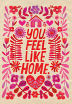 Front of a folk art style greeting card with a colorful house, hearts, and floral designs in pink, red, and purple, featuring the heartfelt message 'YOU FEEL LIKE HOME' for a spouse.