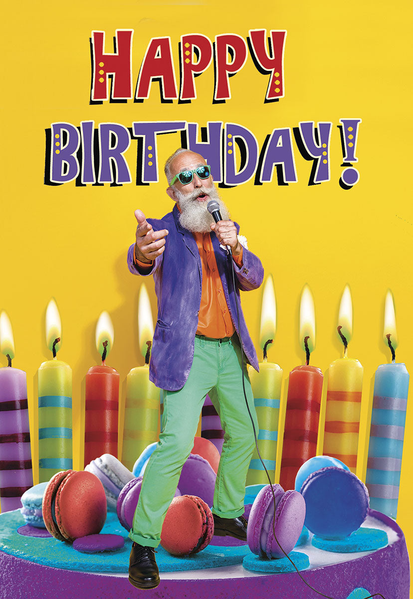 Vibrant yellow funny birthday greeting card featuring a cool, older man with a white beard, sunglasses, purple jacket, and green pants, holding a microphone. He stands on a colorful cake surrounded by lit striped candles and macarons, with 'HAPPY BIRTHDAY!' text above.