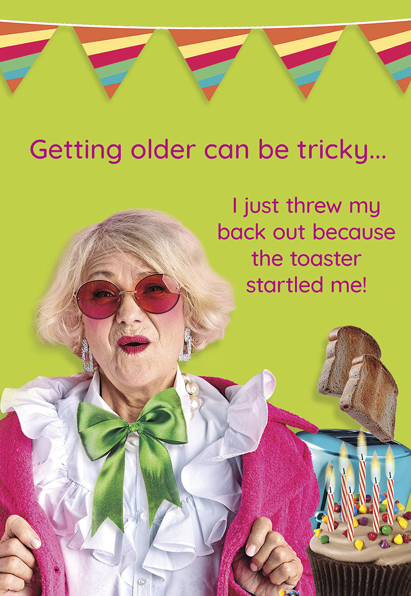 Humorous birthday card front featuring a stylish older woman in large red sunglasses and a pink robe, startled by a toaster popping toast. A cupcake with lit candles sits nearby. Text reads: 'Getting older can be tricky... I just threw my back out because the toaster startled me!' Colorful bunting is at the top.