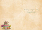 Inside of a vintage-themed greeting card with a blank background and a small cake and flower decoration, displaying the humorous message 'Here's to you getting older... finally. Happy Birthday!'