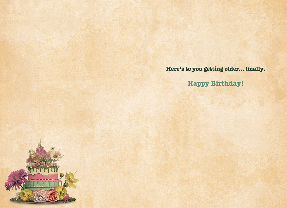 Inside of a vintage-themed greeting card with a blank background and a small cake and flower decoration, displaying the humorous message 'Here's to you getting older... finally. Happy Birthday!'