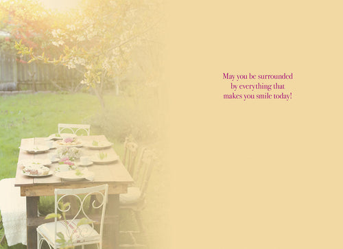 Inside view of a Mother's Day card showing a beautifully set garden table fading into the background, with a heartfelt message 'May you be surrounded by everything that makes you smile today!' in elegant script.