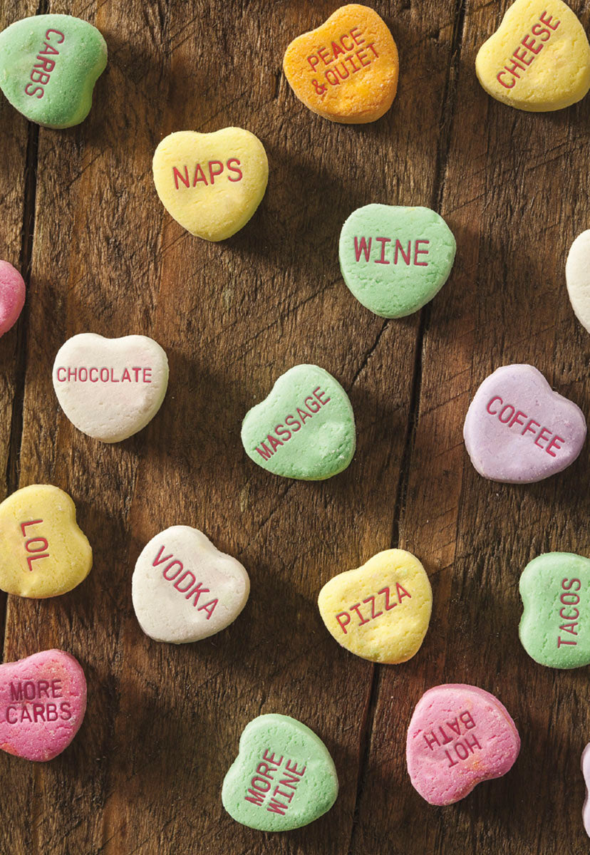 An overhead view of colorful heart-shaped candies with humorous and sarcastic messages like 'Wine', 'Naps', 'Vodka', and 'Pizza' on a wooden surface.