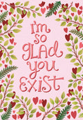 Front of a heartfelt Valentine's Day greeting card featuring the phrase 'I'm so glad you exist' in decorative pink script, framed by green leafy branches with red hearts and berries on a light pink textured background, perfect for a romantic gesture or expressing deep affection to a spouse.