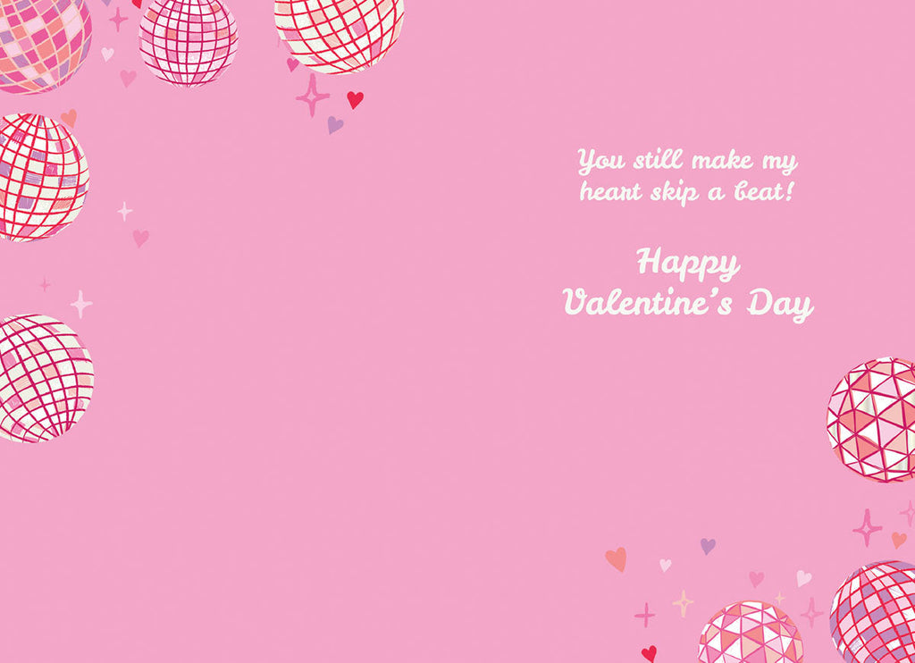 Inside of a playful pink greeting card for a spouse, adorned with scattered disco balls, hearts, and stars, with the romantic message 'You still make my heart skip a beat! Happy Valentine's Day'.